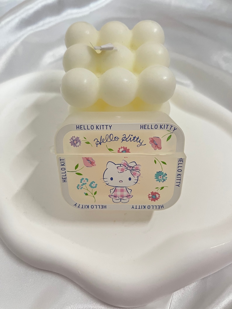 May include: A white Hello Kitty candle with a bubble design. The candle has a Hello Kitty graphic on the front with flowers and the text "Hello Kitty" repeated around the border.