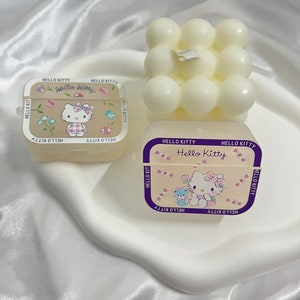 May include: Two Hello Kitty themed candle holders. One is a square with a white Hello Kitty design and the other is a white cube with round bumps.