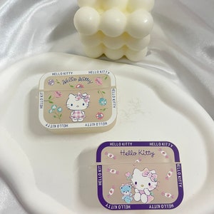 May include: Two Hello Kitty Airpods cases. One case is beige with a floral design and the other is beige with a candy design. Both cases have a purple border and the text "Hello Kitty" on the front.