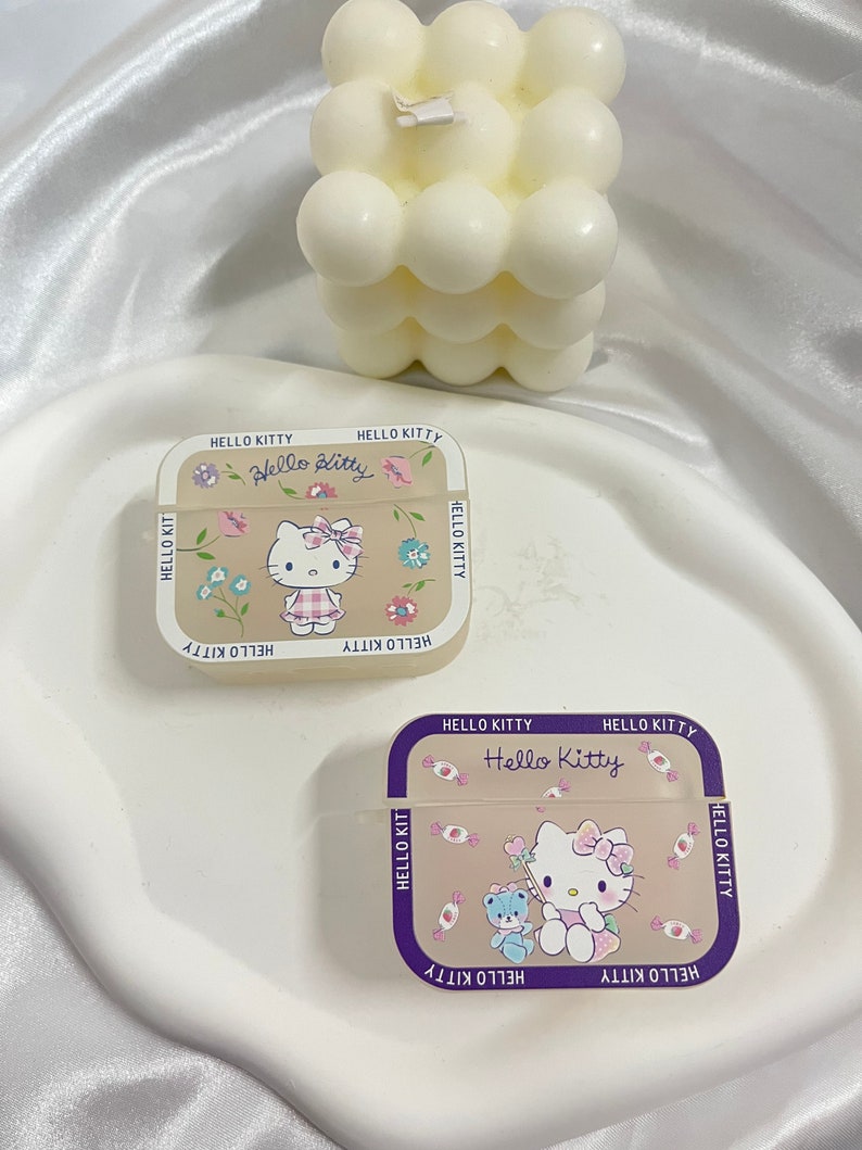 May include: Two Hello Kitty Airpods cases. One case is beige with a floral design and the other is beige with a candy design. Both cases have a purple border and the text "Hello Kitty" on the front.