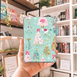 May include: A clear phone case featuring a festive Christmas tree pattern. The design includes various Christmas trees in pink, gold, and teal, adorned with bows and ornaments, set against a light teal background. A seasonal accessory.