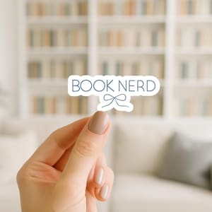 May include: A white sticker with the text "BOOK NERD" in a light blue font, with a small bow graphic. The sticker is held between two fingers against a blurred background of a bookshelf and a couch.