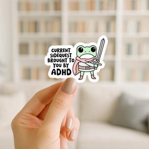May include: A white sticker featuring a cartoon frog warrior holding a sword. The frog wears a pink scarf and cape, with the text "CURRENT SIDEQUEST BROUGHT TO YOU BY ADHD" in black letters.