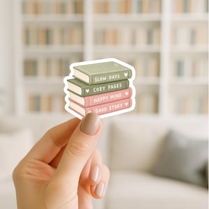 May include: A sticker featuring a stack of four books in shades of green and pink. Each book has a title: "SLOW DAYS", "COZY PAGES", "HAPPY MIND", and "GOOD STORY". Small hearts are next to each title. The sticker is held against a blurred background.