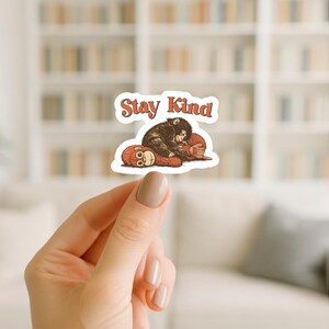 May include: A sticker featuring a brown monkey hugging a red monkey, with the words "Stay Kind" in a retro font. The sticker has a white border and is held up by a hand.