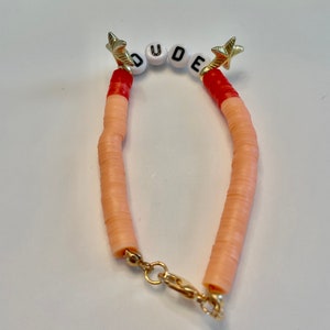 May include: A beaded bracelet with the word "DUDE" spelled out in white letter beads. The bracelet features peach-colored disc beads, red accents, and gold star-shaped charms. A gold clasp secures the bracelet.