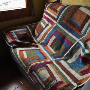 K&ouml;nnte beinhalten: Eine bunte gestrickte Wolldecke mit einem geometrischen Muster aus Quadraten und Rechtecken in Braun-, Gr&uuml;n-, Blau-, Rot- und Wei&szlig;t&ouml;nen. Die Decke liegt auf einem Sofa.