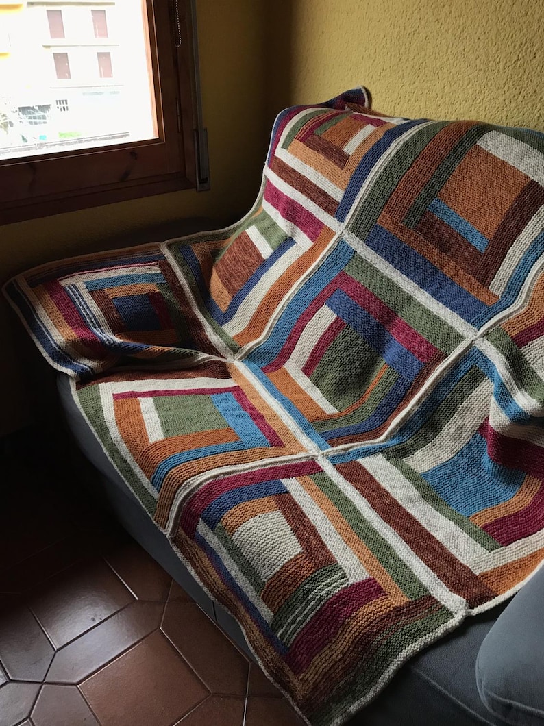 K&ouml;nnte beinhalten: Eine bunte gestrickte Wolldecke mit einem geometrischen Muster aus Quadraten und Rechtecken in Braun-, Gr&uuml;n-, Blau-, Rot- und Wei&szlig;t&ouml;nen. Die Decke liegt auf einem Sofa.