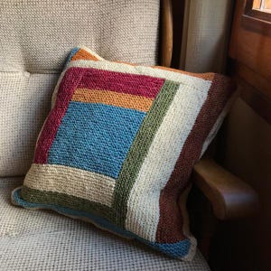 K&ouml;nnte beinhalten: Ein gestricktes Kissen mit einem geometrischen Muster in Beige-, Braun-, Blau-, Gr&uuml;n- und Rott&ouml;nen. Das Kissen liegt auf einem beigen Sessel.
