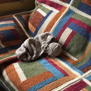 K&ouml;nnte beinhalten: Eine mehrfarbige gestrickte Decke mit einem geometrischen Muster aus Quadraten und Streifen in Braun-, Orange-, Gr&uuml;n-, Blau- und Wei&szlig;t&ouml;nen. Ein Kn&auml;uel Garn und Stricknadeln liegen auf der Decke.