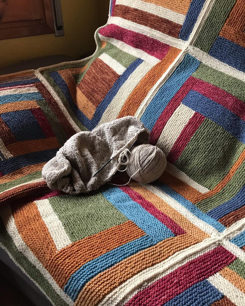K&ouml;nnte beinhalten: Eine mehrfarbige gestrickte Decke mit einem geometrischen Muster aus Quadraten und Streifen in Braun-, Orange-, Gr&uuml;n-, Blau- und Wei&szlig;t&ouml;nen. Ein Kn&auml;uel Garn und Stricknadeln liegen auf der Decke.