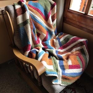 Könnte beinhalten: Eine bunte gestrickte Wolldecke mit Streifen und Quadraten in Blautönen, Grün, Orange, Rot und Weiß. Die Decke liegt über einem Holz-Schaukelstuhl.