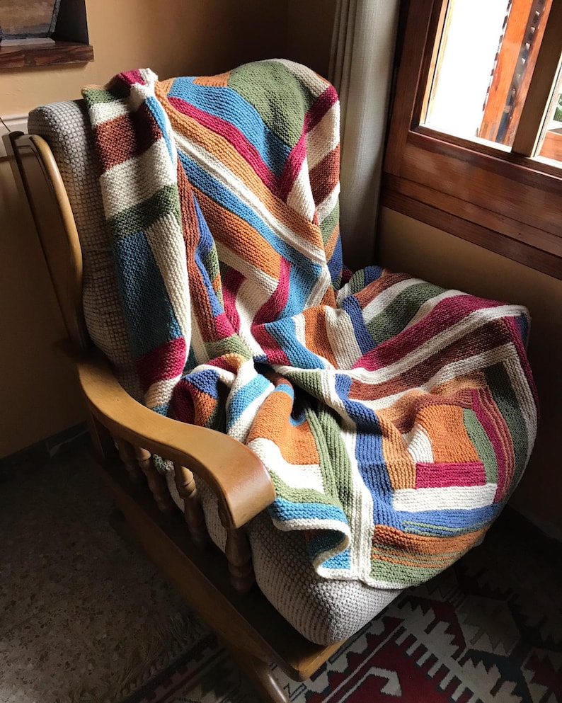 K&ouml;nnte beinhalten: Eine bunte gestrickte Wolldecke mit Streifen und Quadraten in Blaut&ouml;nen, Gr&uuml;n, Orange, Rot und Wei&szlig;. Die Decke liegt &uuml;ber einem Holz-Schaukelstuhl.