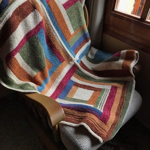 K&ouml;nnte beinhalten: Eine bunte gestrickte Wolldecke mit geometrischem Muster in Braun-, Orange-, Gr&uuml;n-, Blau-, Rot- und Wei&szlig;t&ouml;nen. Die Decke liegt &uuml;ber einem Holz-Schaukelstuhl.