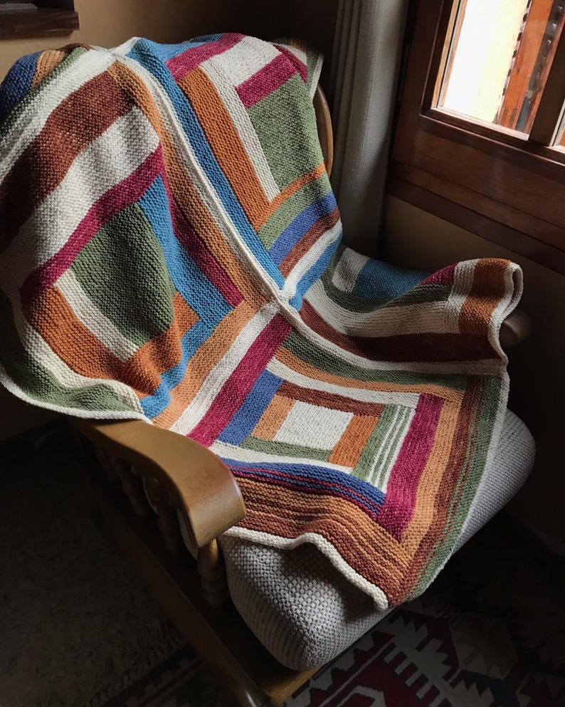 K&ouml;nnte beinhalten: Eine bunte gestrickte Wolldecke mit geometrischem Muster in Braun-, Orange-, Gr&uuml;n-, Blau-, Rot- und Wei&szlig;t&ouml;nen. Die Decke liegt &uuml;ber einem Holz-Schaukelstuhl.