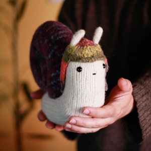 Może przedstawiać: Ręcznie robiona, dziergana zabawka w kształcie ślimaka z kremowym korpusem, bordowo-niebieską muszlą i kolorowym kapeluszem. Ślimak ma małe czarne oczy i wyszywane usta. Zabawka trzymana jest w dłoniach.