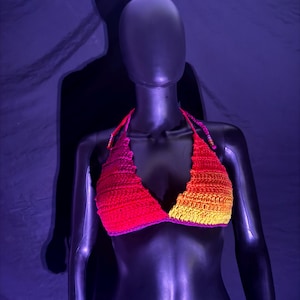 May include: A handmade crochet halter top in a gradient of red, orange, and yellow hues. The top is displayed on a black mannequin against a dark purple backdrop. The crochet work features a scalloped edge and a purple trim.