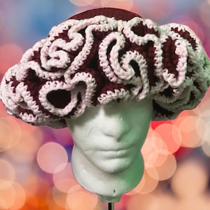 May include: A burgundy and white crocheted hat with a ruffled design. The hat is displayed on a white mannequin head.
