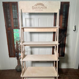 Puede incluir: Estantería de madera clara con múltiples niveles. La sección superior presenta el logotipo "OAKNSMITH WOOD WORKS". Los estantes están soportados por un marco, y la unidad se encuentra sobre un suelo de ladrillo. El fondo incluye una puerta y una ventana blancas.