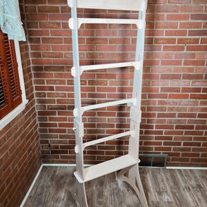 May include: A tall, unfinished wooden display stand with multiple horizontal shelves and a backboard. The stand has a light wood color and is designed for displaying items. It stands on a wooden floor against a brick wall.