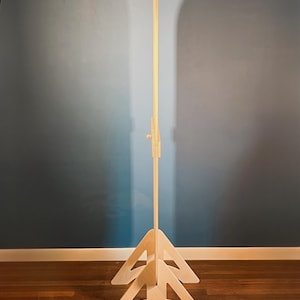 May include: A tall, light-colored wooden floor stand featuring a vertical central pole supported by two triangular, arched base pieces. The structure includes a small, adjustable wooden knob mechanism located on the upper section of the central pole.