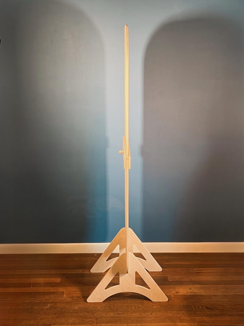 May include: A tall, light-colored wooden floor stand featuring a vertical central pole supported by two triangular, arched base pieces. The structure includes a small, adjustable wooden knob mechanism located on the upper section of the central pole.
