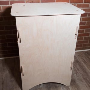 Puede incluir: Un mueble de madera clara con una parte superior rectangular y una base curva. El mueble tiene ensamblajes visibles y está colocado contra una pared de ladrillo y un suelo de madera.