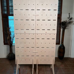May include: A large, unfinished wooden display stand with a light natural wood finish. The stand features multiple rows of evenly spaced holes and slots, with four hinged doors and supporting legs. The design is simple and functional, suitable for various display purposes.