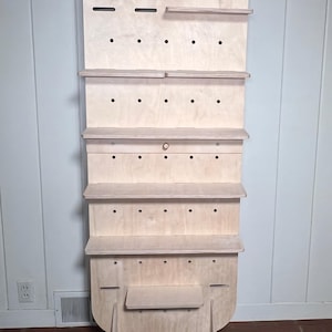 May include: A freestanding wooden shelf unit with a rounded top and multiple shelves. The shelves are made of plywood and have a natural wood finish. The unit has a pegboard design with holes for hanging items. The unit is supported by two angled legs.