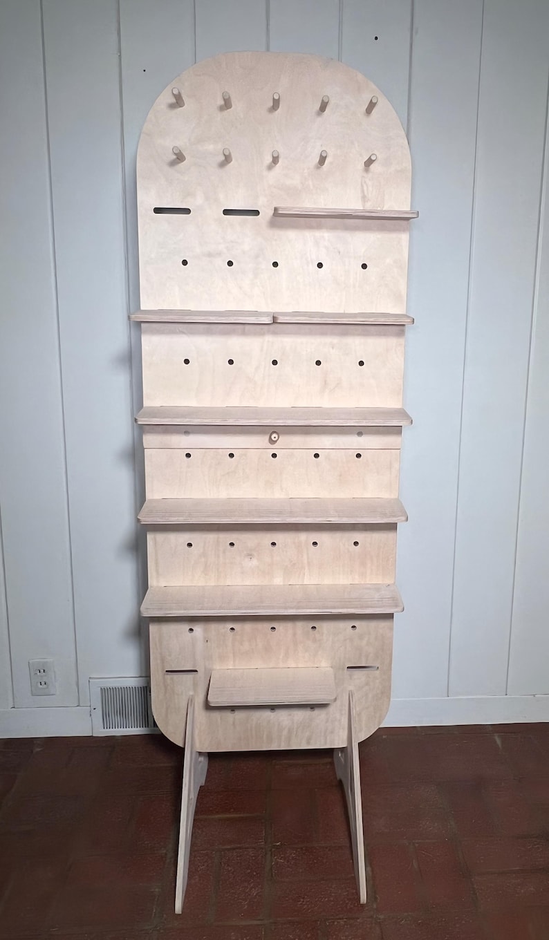 May include: A freestanding wooden shelf unit with a rounded top and multiple shelves. The shelves are made of plywood and have a natural wood finish. The unit has a pegboard design with holes for hanging items. The unit is supported by two angled legs.