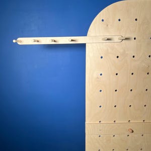 May include: A light-colored wooden pegboard display stand with a rounded top edge, featuring a grid of circular holes. A horizontal wooden bar with five protruding pegs is attached to the upper section of the board, extending to the left side.