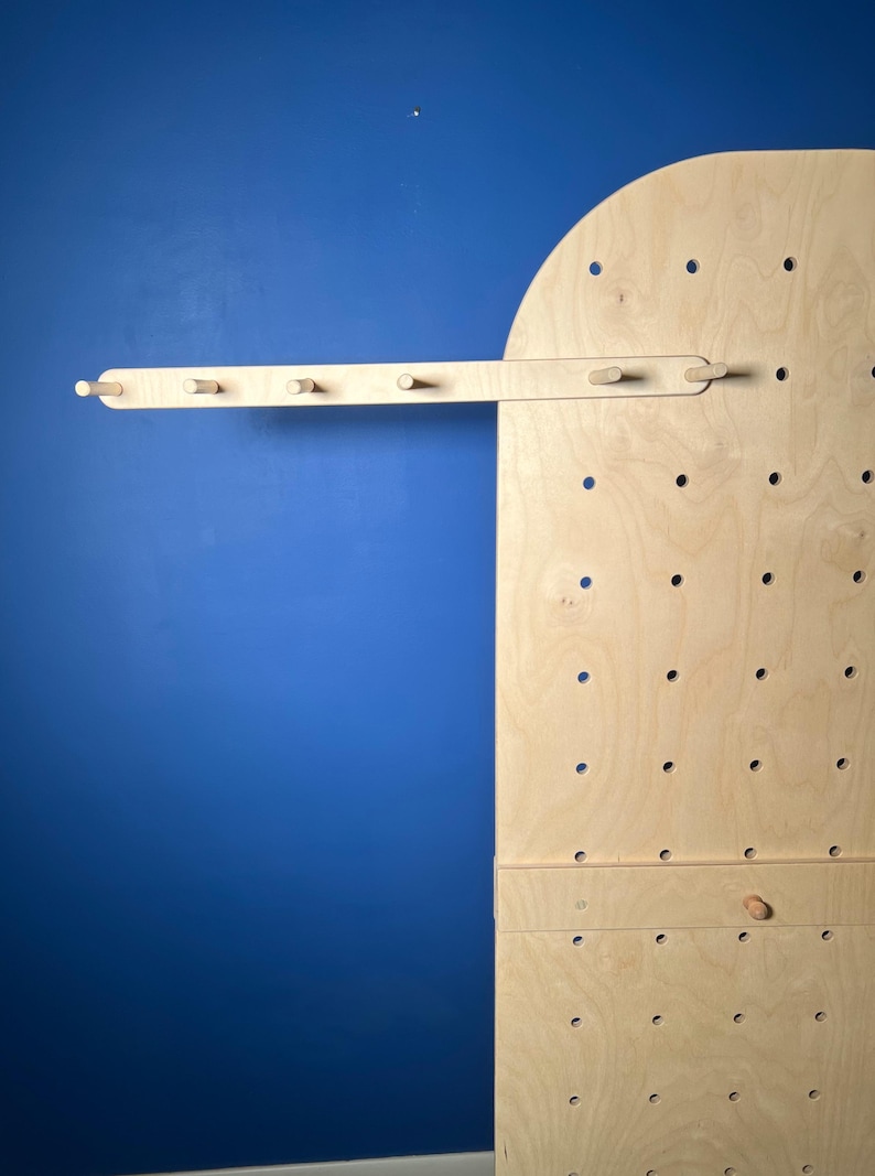 May include: A light-colored wooden pegboard display stand with a rounded top edge, featuring a grid of circular holes. A horizontal wooden bar with five protruding pegs is attached to the upper section of the board, extending to the left side.