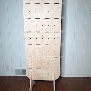 May include: A freestanding, light wood display stand with a rounded top and multiple holes for hanging items. The stand has a simple, modern design and is ideal for showcasing jewellery, accessories, or other small items.
