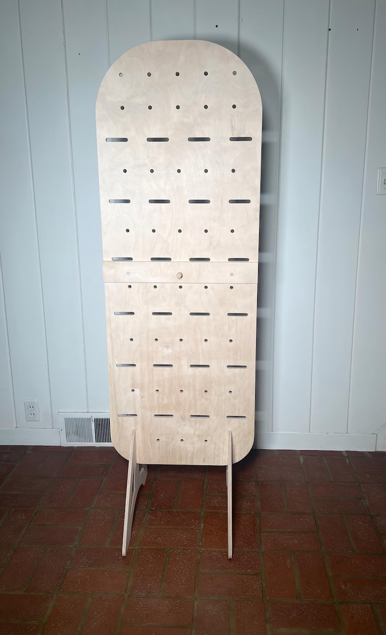 May include: A freestanding, light wood display stand with a rounded top and multiple holes for hanging items. The stand has a simple, modern design and is ideal for showcasing jewellery, accessories, or other small items.