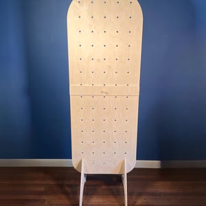 May include: A tall, light-colored wooden pegboard display stand with a rounded top. The board features a grid of evenly spaced circular holes for hanging items and is supported by two triangular wooden feet at the base for stability.