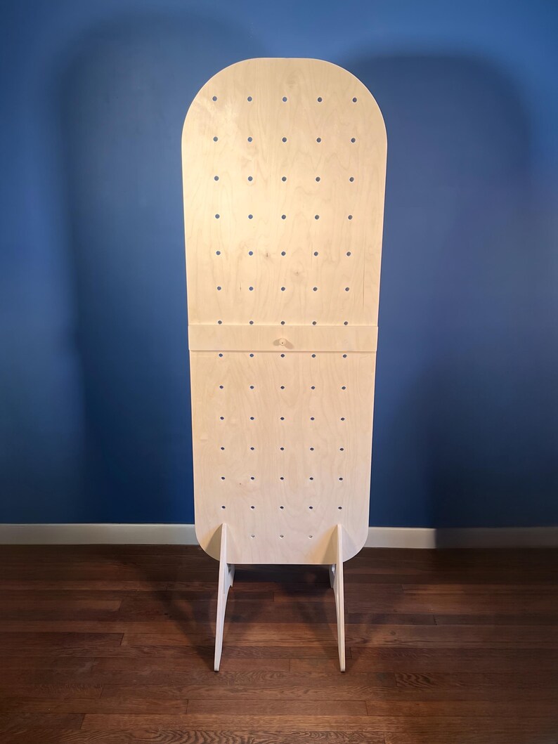 May include: A tall, light-colored wooden pegboard display stand with a rounded top. The board features a grid of evenly spaced circular holes for hanging items and is supported by two triangular wooden feet at the base for stability.