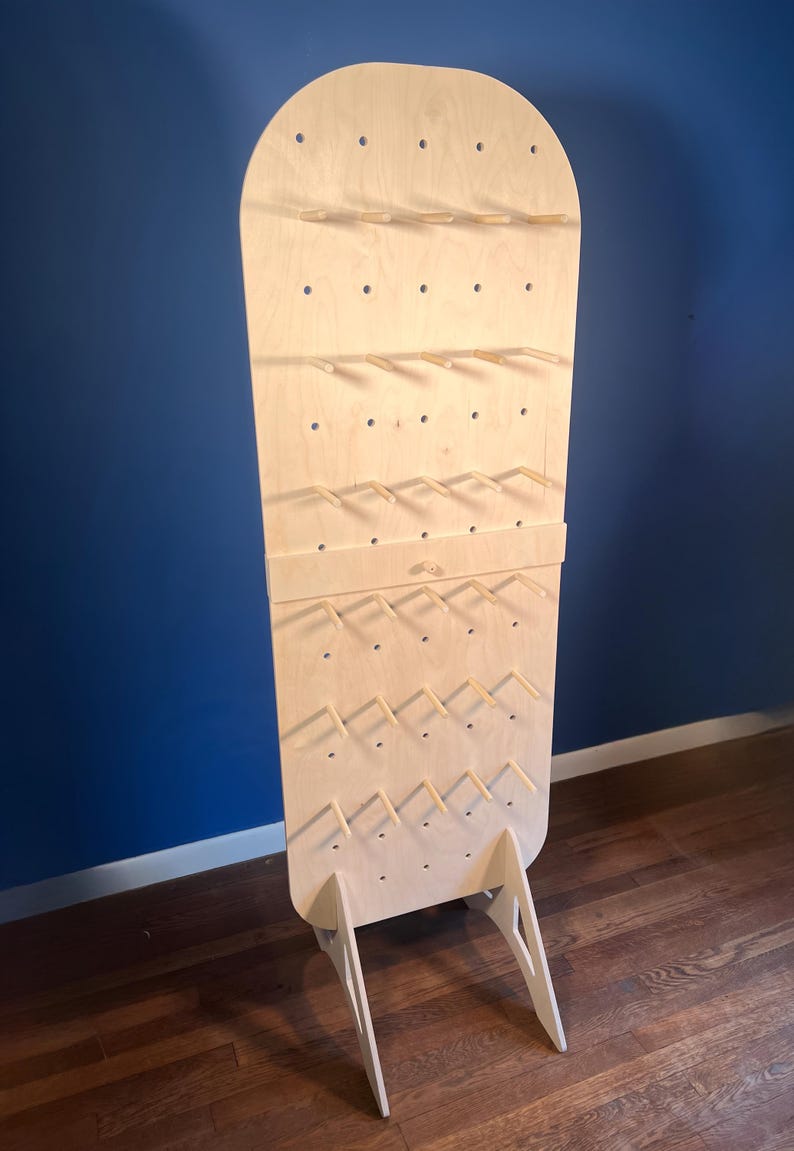 May include: A tall, light-colored wooden display stand with a rounded top, featuring a grid of evenly spaced holes and multiple horizontal wooden pegs extending outward. The structure is supported by two angled wooden legs at the base.