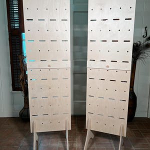 May include: Two light-colored wooden display panels with numerous holes and slots. Each panel has a stand and is designed to hold items. The panels are positioned on a brick floor, suggesting a retail or craft fair setting.