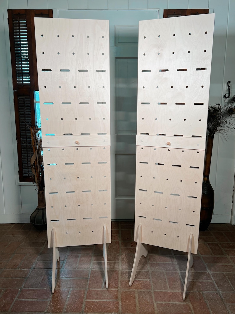 May include: Two light-colored wooden display panels with numerous holes and slots. Each panel has a stand and is designed to hold items. The panels are positioned on a brick floor, suggesting a retail or craft fair setting.