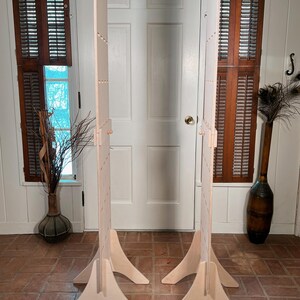 May include: Two unfinished wooden display stands with a light-colored, natural wood finish. Each stand has a tall, rectangular panel supported by a three-legged base. The stands are designed to be freestanding and are suitable for displaying items.