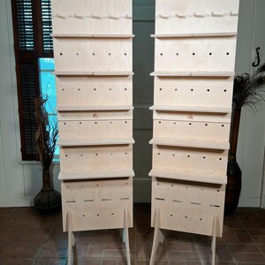 May include: Two unfinished wooden display stands. Each stand has multiple shelves, hooks, and a base with angled legs. The stands are made of light-colored wood and are designed for displaying items.