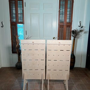 May include: Two unfinished wooden display stands with multiple horizontal slots and small holes. Each stand has a simple, angled leg design. The stands are positioned in front of a white door and brown shutters.