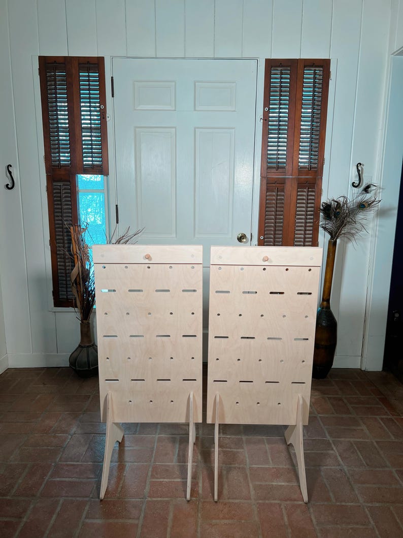 May include: Two unfinished wooden display stands with multiple horizontal slots and small holes. Each stand has a simple, angled leg design. The stands are positioned in front of a white door and brown shutters.