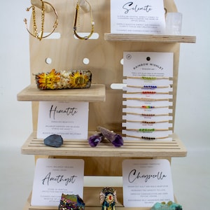 May include: A wooden display stand with shelves showcasing various crystals and jewelry. The crystals are labeled with their names and properties, such as amethyst, selenite, hematite, and chrysocolla. The jewelry includes a rainbow wishlet bracelet set and gold bracelets.