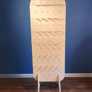 May include: A tall, light-colored wooden display board with a rounded top and a flat base. The board features a grid of evenly spaced circular holes, with several small wooden pegs inserted into the holes to hold items. It stands on two legs.