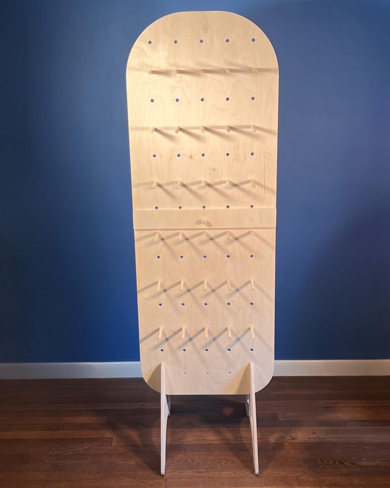May include: A tall, light-colored wooden display board with a rounded top and a flat base. The board features a grid of evenly spaced circular holes, with several small wooden pegs inserted into the holes to hold items. It stands on two legs.