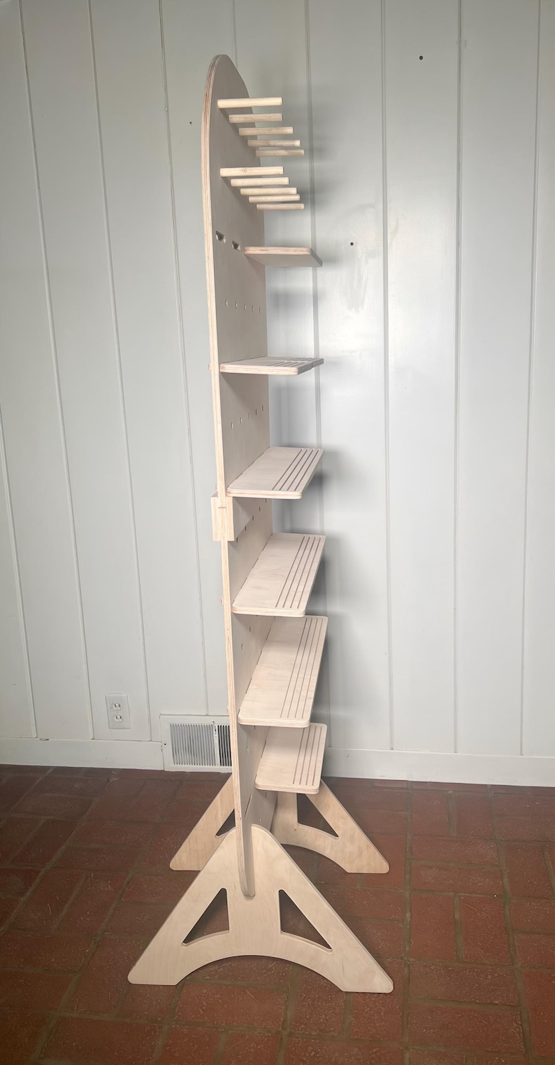May include: A freestanding wooden shelf unit with five shelves and a pegboard back. The shelf unit has a triangular base and is made of light-coloured wood.