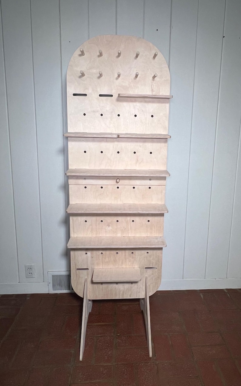 May include: A tall, freestanding wooden shelf unit with a rounded top and multiple shelves. The shelves are made of light-coloured wood and have a series of small holes drilled into them. The unit is supported by two angled legs.