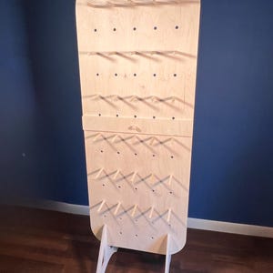 May include: A tall, light-colored wooden pegboard display stand with a rounded top. The vertical board features a grid of circular holes with several horizontal wooden pegs inserted, supported by two angled wooden legs at the base.