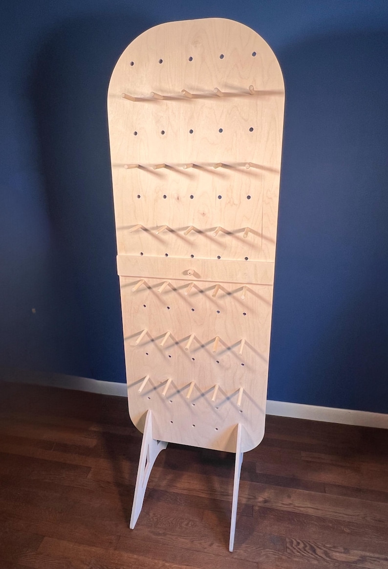 May include: A tall, light-colored wooden pegboard display stand with a rounded top. The vertical board features a grid of circular holes with several horizontal wooden pegs inserted, supported by two angled wooden legs at the base.